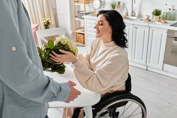 Μια ανάπηρη γυναίκα σε αναπηρικό καροτσάκι που κρατάει ένα ζωντανό μπουκέτο λουλούδια, περιτριγυρισμένη από αγάπη στην κουζίνα του σπιτιού της. - Φωτογραφία, εικόνα
