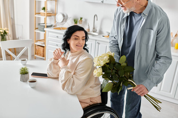Un homme tenant un bouquet, se tient amoureusement à côté de sa femme alors qu'elle est assise dans un fauteuil roulant dans leur cuisine à la maison. - Photo, image