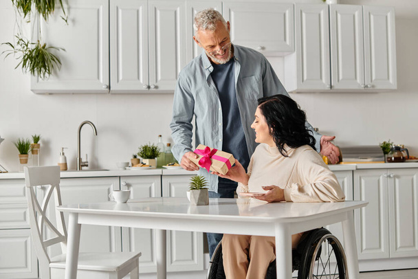 Un uomo porge teneramente un regalo a una donna disabile su una sedia a rotelle, nella loro cucina di casa.. - Foto, immagini