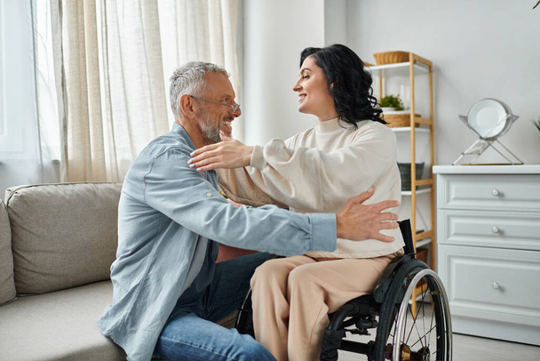 Μια γυναίκα σε αναπηρικό καροτσάκι αγκαλιάζει τον άντρα της, δείχνοντας αγάπη και υποστήριξη στο σαλόνι τους.. - Φωτογραφία, εικόνα