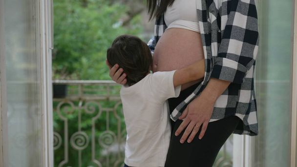 Niño cariñoso besándose y abrazando el vientre de la madre en el tercer trimestre del embarazo mostrando afecto hacia el hermano bebé nonato - Foto, imagen