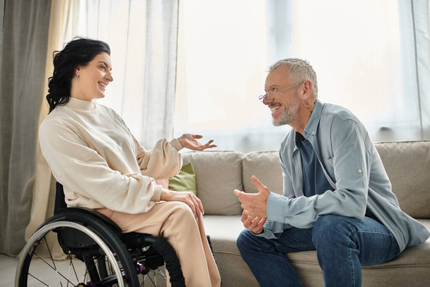 Ein Mann unterhält sich in gemütlicher Wohnzimmeratmosphäre mit einer behinderten Frau im Rollstuhl. - Foto, Bild