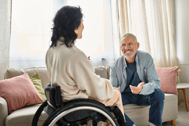 Жінка в інвалідному візку бере участь у розмові з чоловіком у їх затишній обстановці вітальні. - Фото, зображення