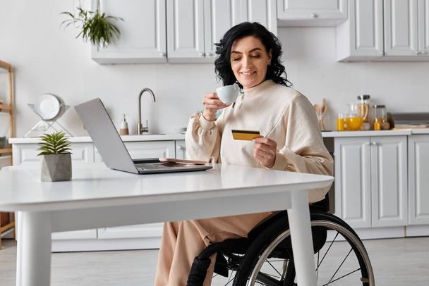 Una mujer discapacitada en silla de ruedas sostiene una taza de café mientras mira una tarjeta de crédito, trabajando remotamente desde su cocina. - Foto, imagen