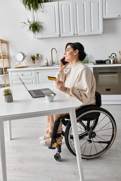 Женщина-инвалид в инвалидном кресле разговаривает по мобильному телефону, работая удаленно со своей кухни. - Фото, изображение