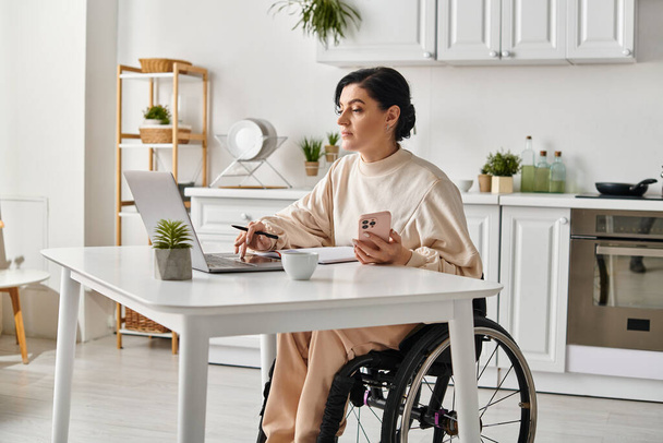 Una mujer discapacitada en una silla de ruedas utilizando un portátil en su casa en su cocina para el trabajo remoto. - Foto, imagen
