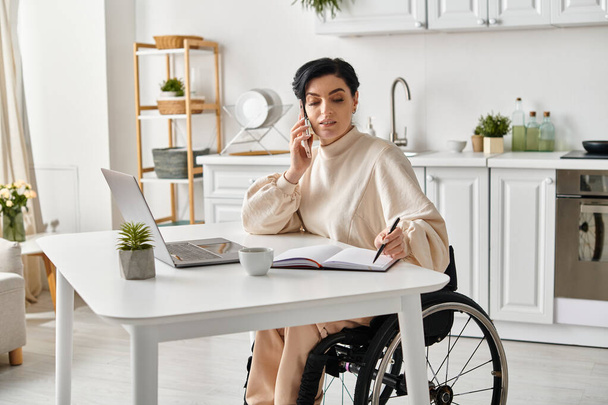 Μια γυναίκα σε αναπηρική καρέκλα εργάζεται εξ αποστάσεως σε ένα τραπέζι με ένα φορητό υπολογιστή στην κουζίνα της. - Φωτογραφία, εικόνα