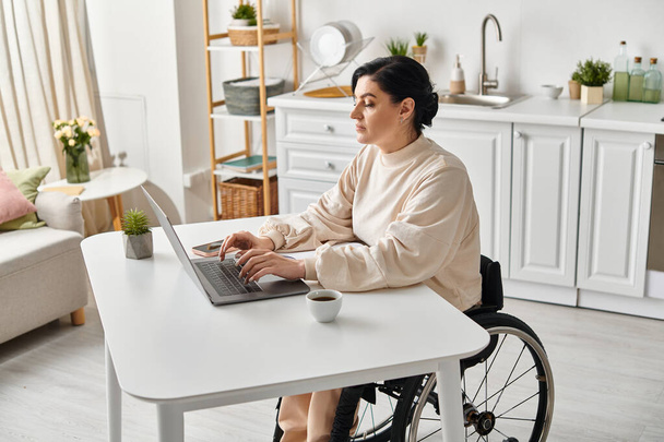 Une femme handicapée en fauteuil roulant travaille à distance sur son ordinateur portable dans la cuisine, mettant en valeur l'indépendance numérique. - Photo, image
