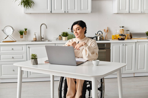 Kobieta na wózku inwalidzkim pracuje zdalnie przy stole kuchennym, używając laptopa, aby utrzymać łączność i produktywność. - Zdjęcie, obraz