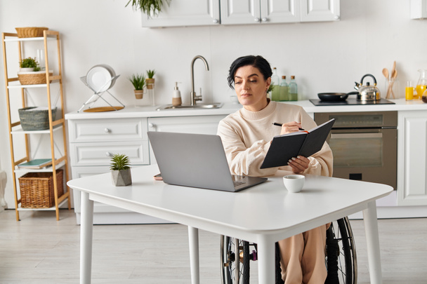 Egy mozgássérült nő tolószékben ül egy konyhaasztalnál, eltökélten és összpontosítva dolgozik a laptopján.. - Fotó, kép
