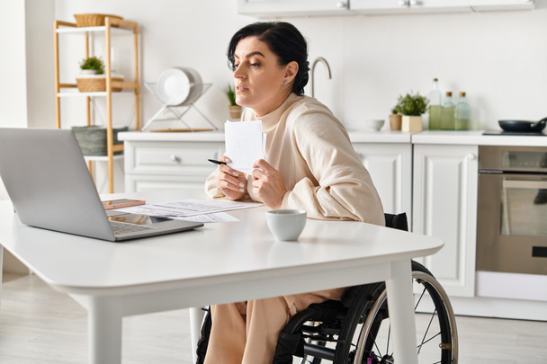 Eine behinderte Frau im Rollstuhl arbeitet von ihrer Küche aus an ihrem Laptop. - Foto, Bild