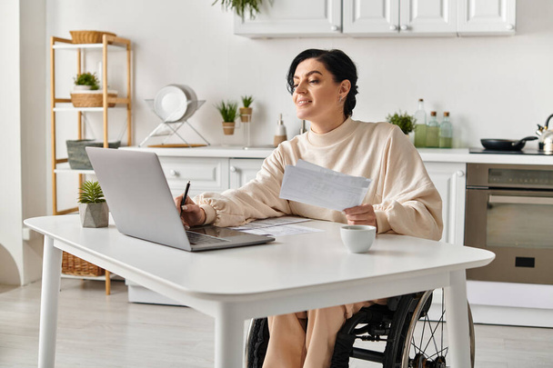 Uma mulher deficiente em uma cadeira de rodas trabalhando remotamente de sua cozinha, usando um computador portátil em uma mesa. - Foto, Imagem