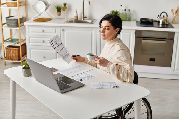 Egy tolószékes nő dolgozik a laptopján, papírokkal körülvéve, kényelmes konyhai környezetben.. - Fotó, kép