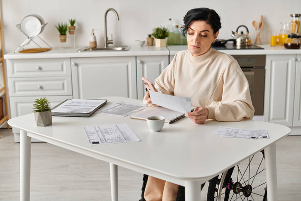 Eine behinderte Frau im Rollstuhl sitzt an einem Küchentisch mit Papieren und einer Tasse Kaffee und konzentriert sich auf Aufgaben aus der Ferne.. - Foto, Bild