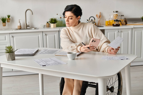 Egy tolószékes mozgássérült nő kávét kortyolgat egy asztalnál a konyhájában, miközben távolról dolgozik.. - Fotó, kép