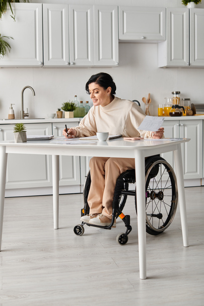 Μια ανάπηρη γυναίκα σε αναπηρικό καροτσάκι δουλεύει σε τραπέζι κουζίνας.. - Φωτογραφία, εικόνα