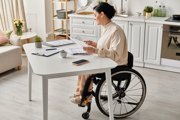 Μια ανάπηρη γυναίκα σε αναπηρικό καροτσάκι που εργάζεται εξ αποστάσεως σε τραπέζι κουζίνας. - Φωτογραφία, εικόνα