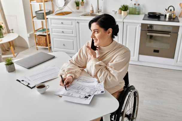 Μια ανάπηρος γυναίκα σε αναπηρική καρέκλα εργάζεται εξ αποστάσεως από την κουζίνα της, κάθεται σε ένα τραπέζι. - Φωτογραφία, εικόνα