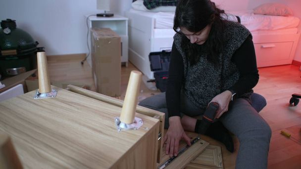 Mujer entusiasta del bricolaje instalando mesita de noche en un nuevo hogar, empleando una máquina de perforación para el ensamblaje de muebles, capturando la esencia de la configuración del hogar y la iniciativa personal - Foto, Imagen