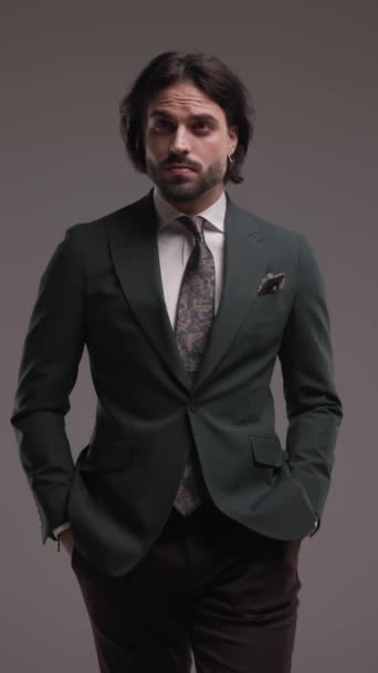 ελκυστική κομψό επιχειρηματία φορώντας κοστούμι και γραβάτα, κοιτάζοντας γύρω, βήμα προς τα εμπρός με τα χέρια στις τσέπες σε γκρι φόντο - Πλάνα, βίντεο
