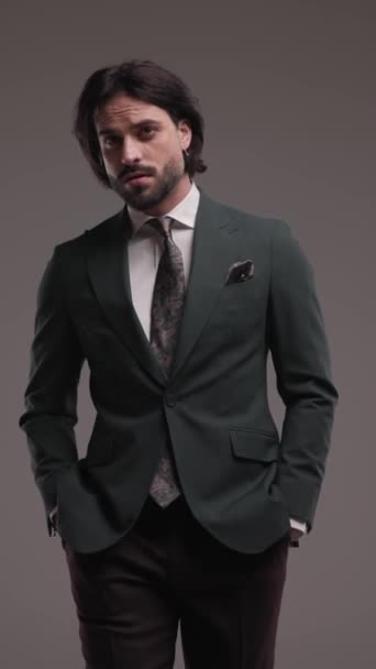 sexy unshaved man walking with hands in pockets, adjusting suit, rubbing palms, fixing beard and looking to side in front of grey background - Footage, Video