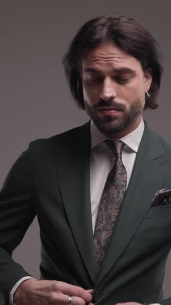 δροσερό γενειοφόρος άνθρωπος προσαρμογή κομψό κοστούμι και γραβάτα, κοιτάζοντας προς τα πλάγια και με αυτοπεποίθηση περπάτημα με τα χέρια στις τσέπες σε γκρι φόντο - Πλάνα, βίντεο