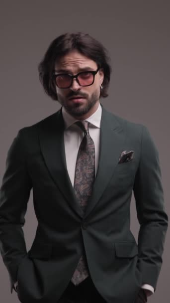 verticale video van zelfverzekerde zakenman met een zonnebril stapt met de handen in zakken, arrangeert pak en kijkt naar opzij, houdt hand in hand en is cool op grijze achtergrond - Video