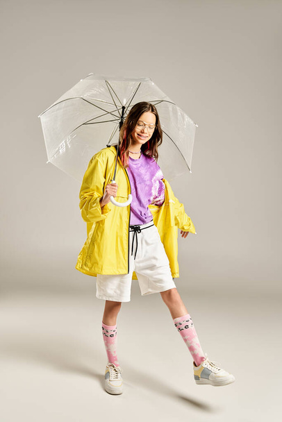 Ένα κομψό έφηβο κορίτσι σε ένα κίτρινο αδιάβροχο κρατά με χαρά μια ομπρέλα, ακτινοβολεί ζωντανή ενέργεια και θετικότητα. - Φωτογραφία, εικόνα