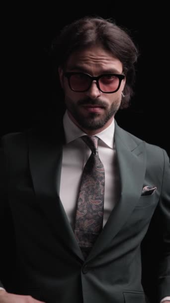atractivo hombre de negocios elegante con gafas de sol mirando hacia abajo, ajustando el traje, tomándose de la mano en los bolsillos y mirando hacia un lado delante del fondo negro - Imágenes, Vídeo