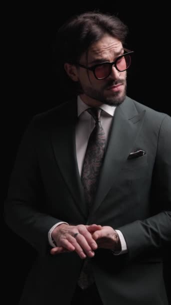 elegante chico de moda con gafas de sol mirando a un lado, la organización de traje y corbata, siendo confiado y posando sobre fondo negro - Imágenes, Vídeo