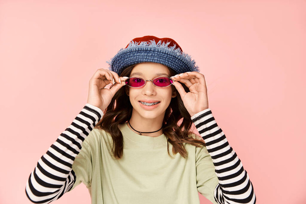 Een stijlvol tiener meisje slaat een pose in levendige kleding, het dragen van een zonnebril en een hoed. - Foto, afbeelding