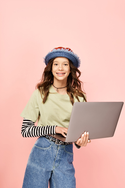 Stílusos tinédzser lány farmerban és kalapban, magabiztosan tartja a laptopot. - Fotó, kép
