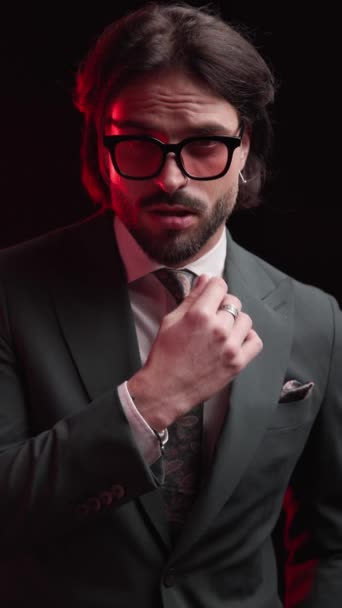 sensuele jonge man in pak met een zonnebril aanraken lippen in een verleidelijke manier, op zoek naar kant en hand in hand te houden in zakken op zwarte achtergrond met rode lichten op - Video