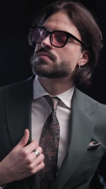 close up video of sensual young man with sunglasses adjusting suit, moving and posing, looking to side in front of black background - Footage, Video