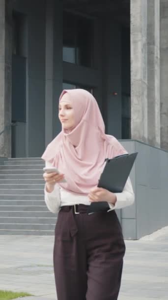 Tela vertical: Vista de baixo ângulo da bela mulher muçulmana que está perto do edifício de escritórios moderno com smartphone nas mãos. Jovem senhora de negócios em desgaste formal e hijab verificando e-mail de trabalho - Filmagem, Vídeo