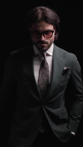 σέξι αξύριστος επιχειρηματίας με κομψό κοστούμι που κρατάει τα χέρια στις τσέπες, περπατά με αυτοπεποίθηση και χαμογελά σε μαύρο φόντο - Πλάνα, βίντεο