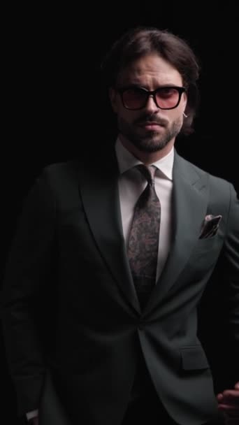 κάθετο βίντεο με αυτοπεποίθηση επιχειρηματίας με κοστούμι περπάτημα με το χέρι στις τσέπες, κοιτάζοντας προς τα κάτω και προς τα εμπρός, προσαρμογή κοστούμι και γυαλιά ηλίου και χαμογελώντας σε μαύρο φόντο - Πλάνα, βίντεο