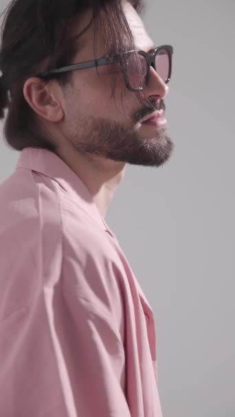 ver video de sexy hombre sin afeitar con gafas de sol en camisa rosa mirando a un lado y tocando la barba sobre fondo gris - Metraje, vídeo