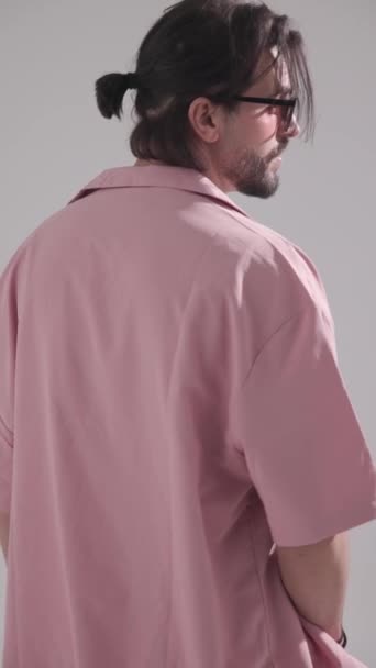 back view of casual fashion man in pink shirt wearing sunglasses moving to side view, rubbing palms and adjusting hair on grey background - Footage, Video