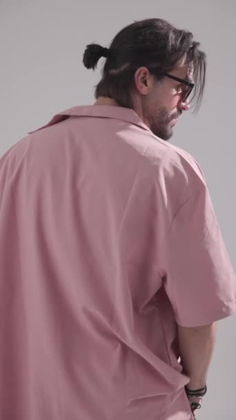 vista trasera del hombre barbudo con pelo largo en camisa rosa con gafas de sol ajustando la ropa y alejándose sobre fondo gris - Metraje, vídeo