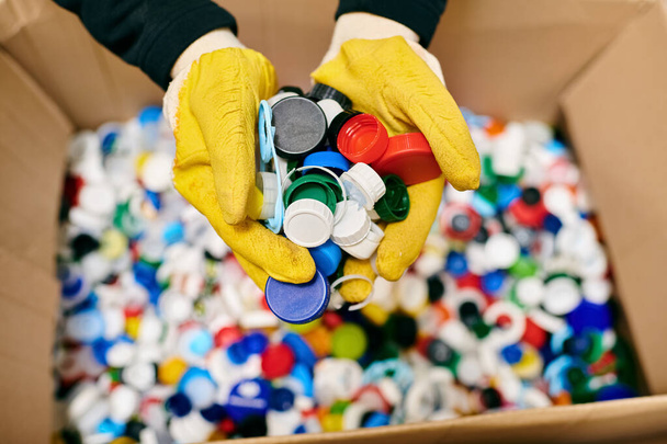 A young volunteer in yellow gloves carefully holding a bunch of colorful bottle caps while sorting trash with eco-conscious peers. - Foto, Bild