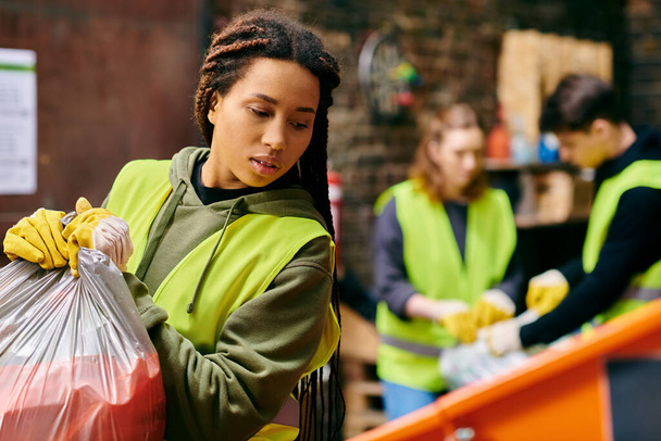 A young woman in a green vest and gloves holding a bag of garbage, participating in a trash sorting activity with other eco-conscious volunteers. - Foto, imagen