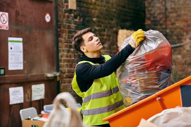 A young man in a yellow vest holds a bag of garbage while volunteering to sort trash with others. - 写真・画像