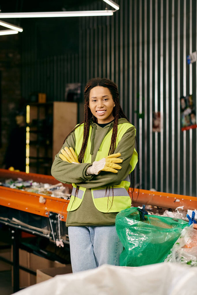 Young volunteer in gloves and safety vest stands with crossed arms, sorting waste in a warehouse. - Photo, Image