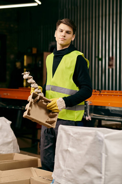 A young volunteer in gloves and safety vest holding a box in a warehouse while sorting waste. - Zdjęcie, obraz