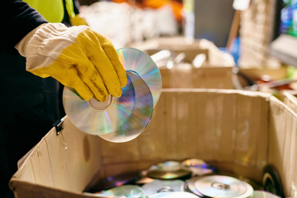 A person in yellow gloves carefully holds a CD inside a box, sorting waste for a cleaner environment. - 写真・画像