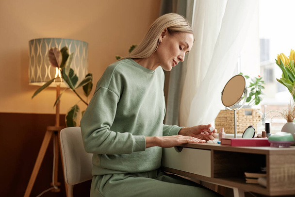 Zijaanzicht portret van blonde volwassen vrouw doen manicure aan ijdelheid tafel door venster kopiëren ruimte - Foto, afbeelding
