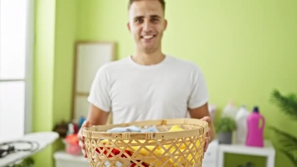Un joven sonriente sostiene una cesta de lavandería en una habitación verde brillante con artículos de lavado en el fondo. - Metraje, vídeo