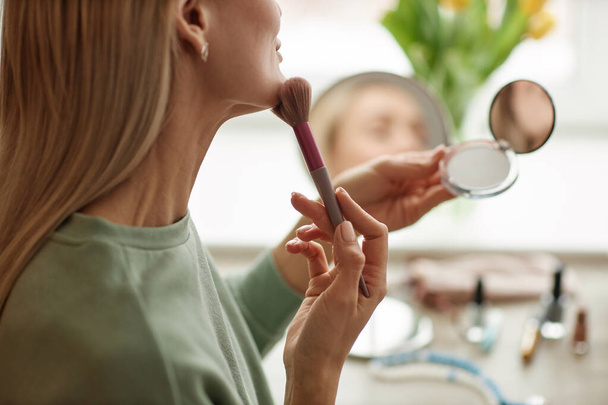 Boční pohled detailní záběr dospělé ženy dělá make-up při pohledu do kompaktního zrcadla podle okna kopírovat prostor - Fotografie, Obrázek