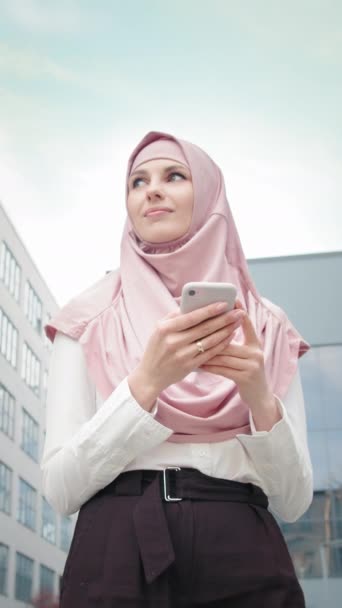 Pantalla vertical: dama musulmana de negocios con móvil de pie cerca del centro de oficinas. Vista de ángulo bajo de la hermosa mujer musulmana de pie cerca del moderno edificio de oficinas con teléfono inteligente en las manos - Imágenes, Vídeo